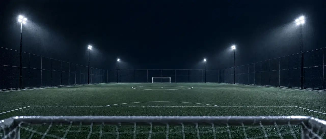 A long, empty floodlit turf field seen from behind a goal
